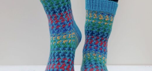 Person wearing colourful knitted socks with intricate patterns of blue, red, green, and yellow, standing on a white wooden surface against a neutral background.