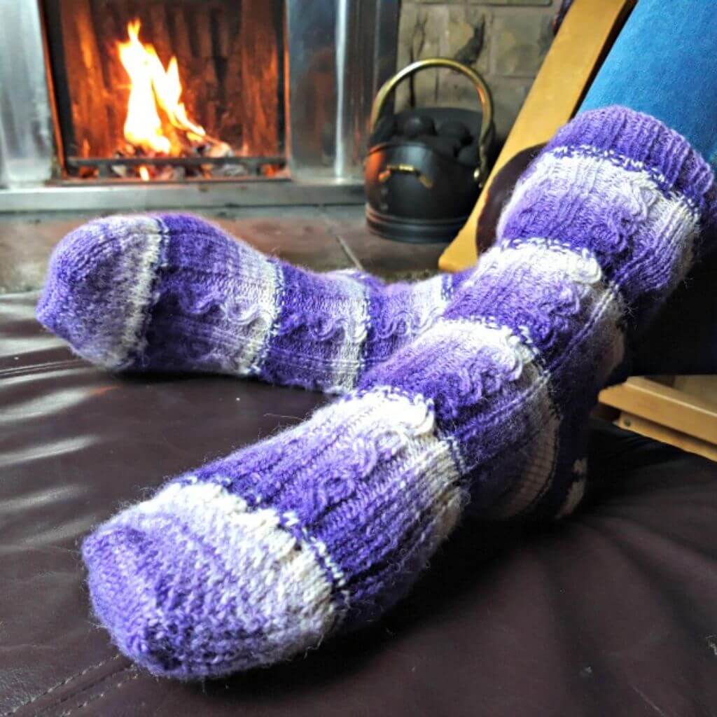 Cozy purple knitted socks rest on a leather footstool in front of a glowing fireplace, creating a warm and relaxed atmosphere. Winwick Mum | Sanctuary Socks