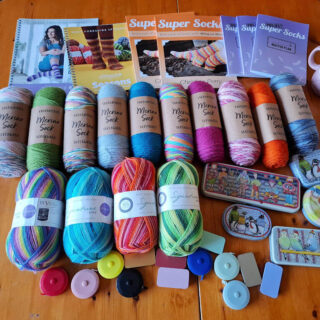 Assorted colourful yarns and knitting supplies on a wooden table. Includes West Yorkshire Spinners and Wool Warehouse yarns, knitting book, tape measures, and mugs with playful designs, creating a cozy, crafty vibe.