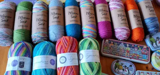 Assorted colourful yarns and knitting supplies on a wooden table. Includes West Yorkshire Spinners and Wool Warehouse yarns, knitting book, tape measures, and mugs with playful designs, creating a cozy, crafty vibe.