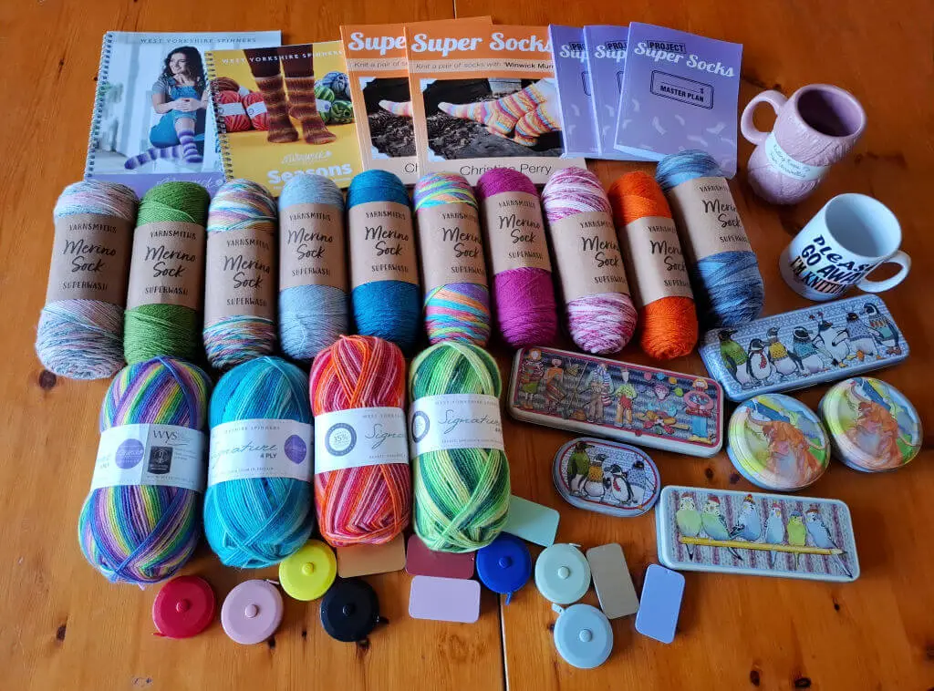 Assorted colourful yarns and knitting supplies on a wooden table. Includes West Yorkshire Spinners and Wool Warehouse yarns, knitting book, tape measures, and mugs with playful designs, creating a cozy, crafty vibe.