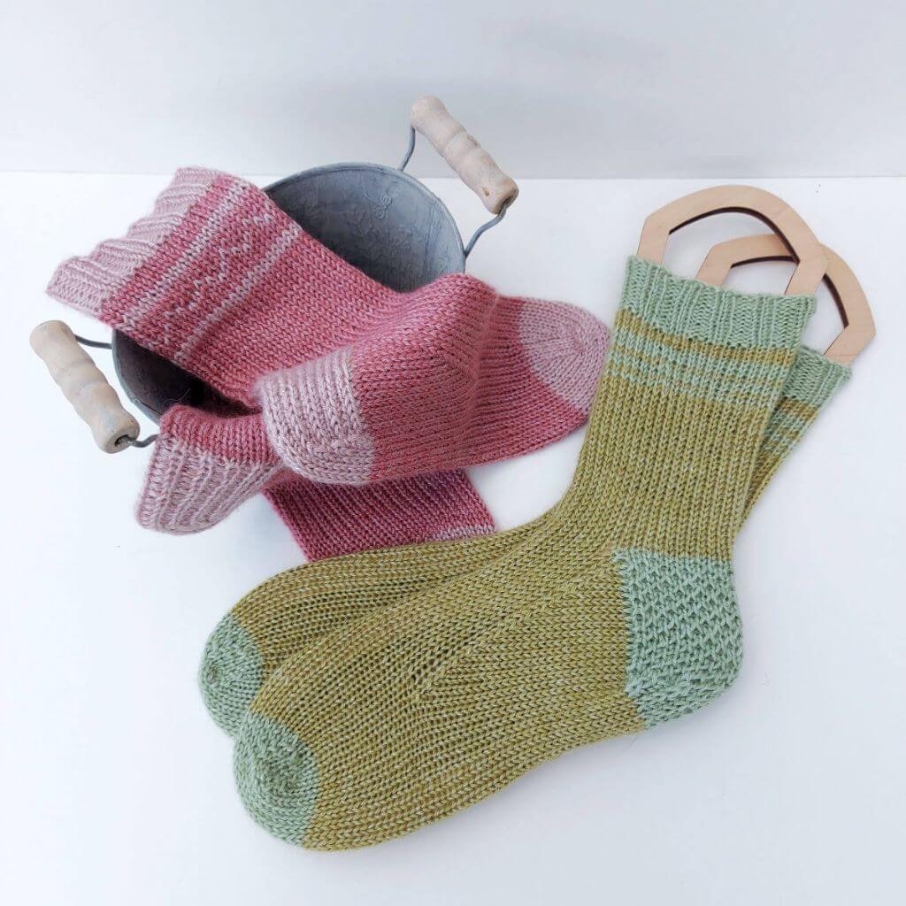 Two pairs of socks arranged on a white background with a small metal bucket. There is a pink pair draped across the bucket and a green pair on sock blockers. Winwick Mum | Sofa Snuggle Socks