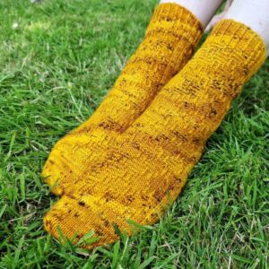 Knitted yellow socks with brown speckles on feet, resting on vibrant green grass. The socks have a cozy and handcrafted appearance.