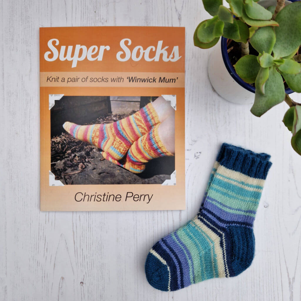 Knitting book "Super Socks" by Christine Perry on a white surface with colorful sock image. Nearby, a handmade striped sock and a green potted plant. Winwick Mum | Super Socks book | beginner sock knitting