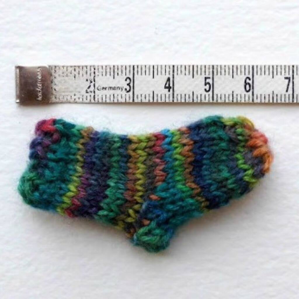 A small, multicolored knitted sock is placed beside a measuring tape on a white background. The sock is approximately 5 centimeters long. Winwick Mum | Teeny Tiny Socks