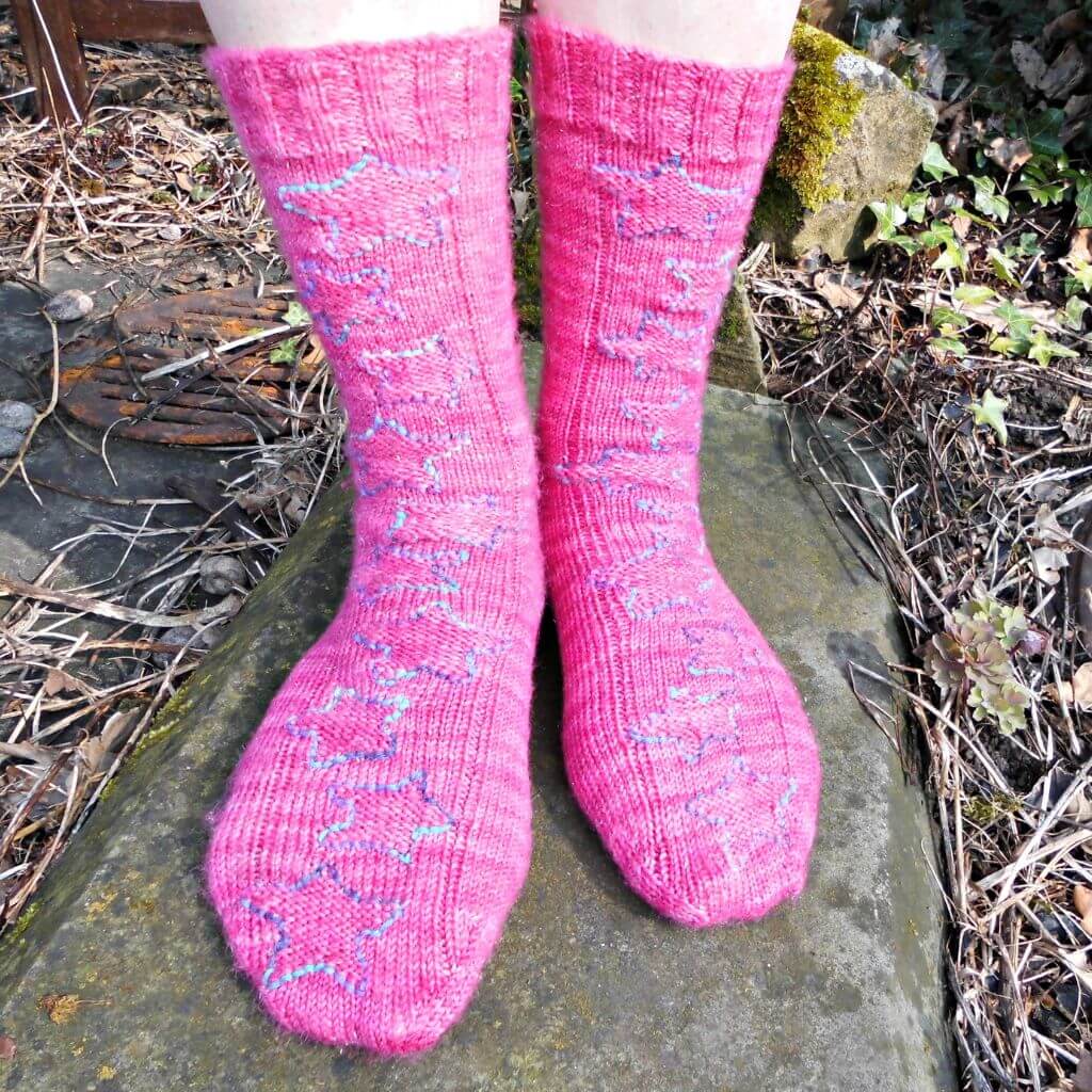 Bright pink socks with a star pattern in blue and green are worn on feet standing on a mossy stone path, surrounded by dry leaves and plants. Winwick Mum | Twinkle Star Socks