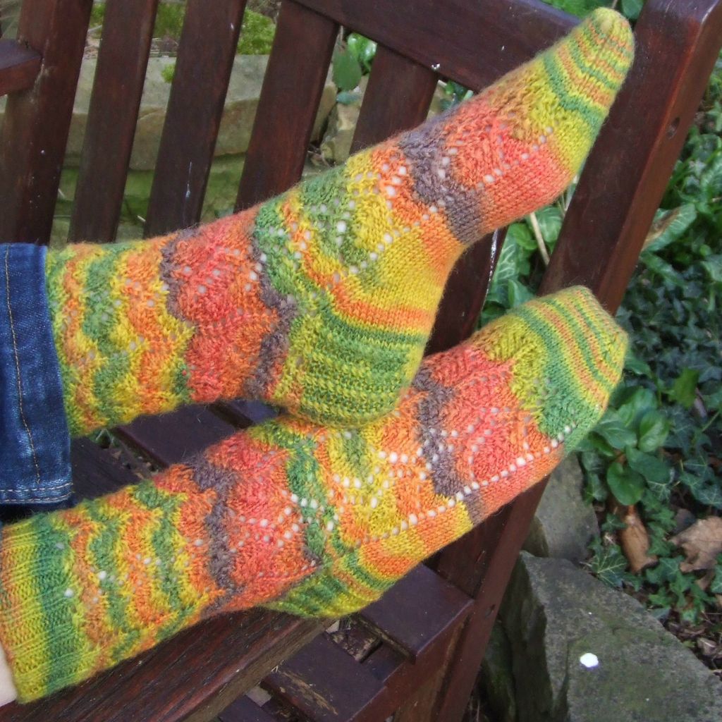 Colourful hand-knitted socks with striped patterns in orange, green, and yellow hues rest on a wooden bench, surrounded by vibrant garden foliage. Winwick Mum | Watercress Leaves Socks