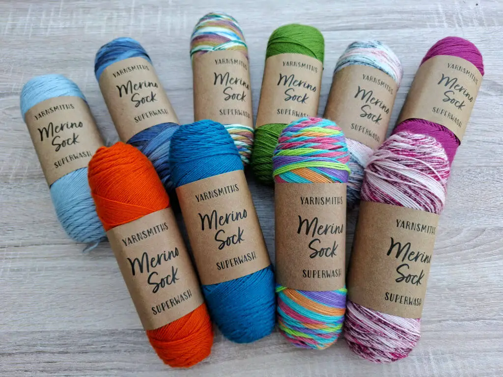 Ten balls of colourful Yarnsmiths Merino Sock Superwash yarn displayed on a wooden surface. Colours include blue, orange, green, and multicolour.