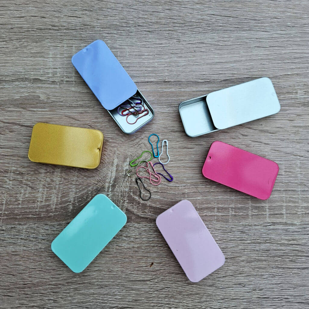 Wooden surface with colourful slide-top tins and hat-shaped markers. Tins in blue, pink, gold, teal, and purple encircle multicoloured markers.