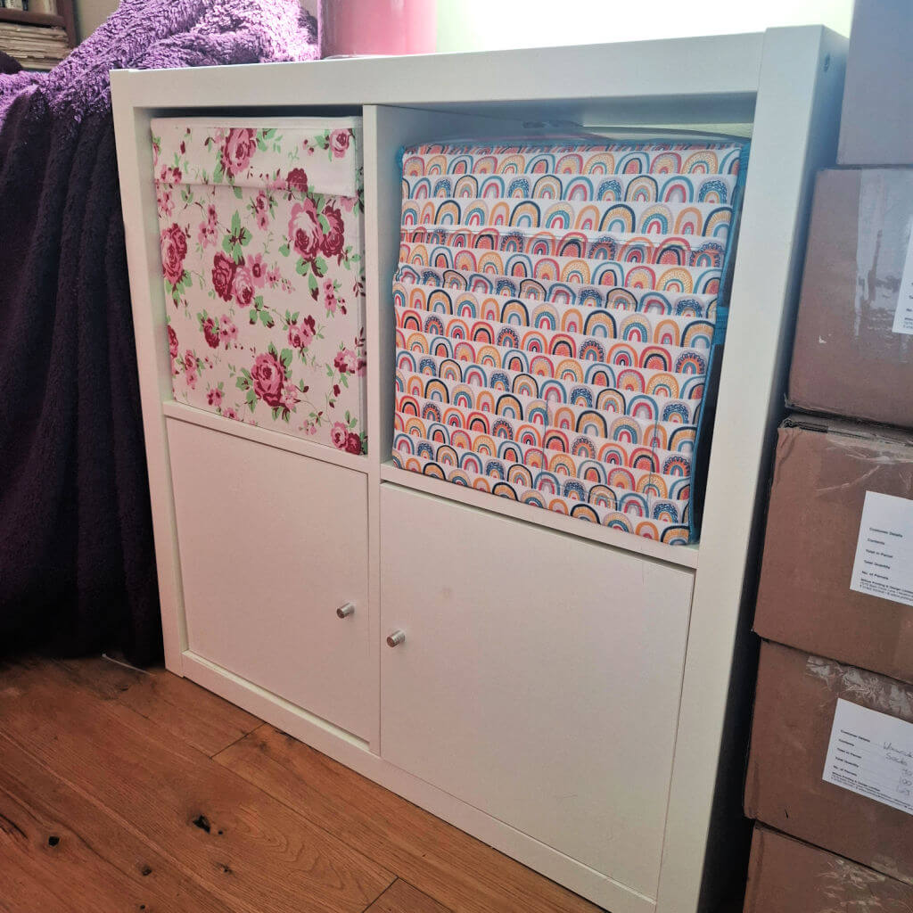 A white IKEA Kallax storage unit on wooden floor holds a floral-patterned box and a rainbow-patterned box. Beside it are brown cardboard boxes. The scene feels organized and cozy.