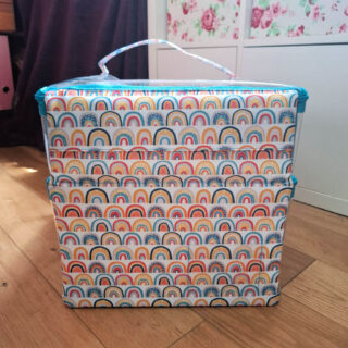A colourful Scubl storage box with a rainbow pattern sits on a wooden floor, next to an IKEA Kallax unit. The box has a handle and conveys a cheerful vibe. Winwick Mum Scubl box review