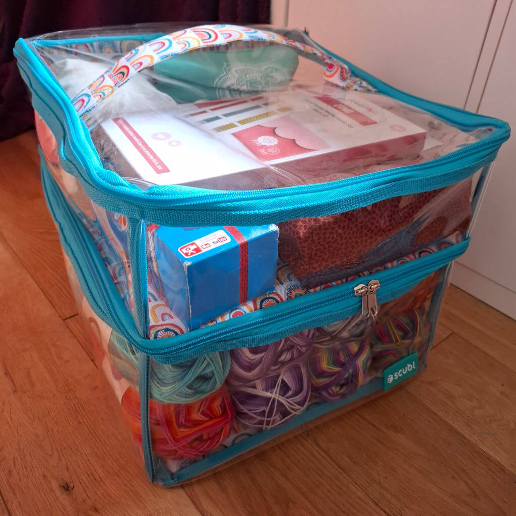 A Scubl storage box with blue zippers on a wooden floor, filled with colourful yarn and accessories, conveying a creative, organized vibe.