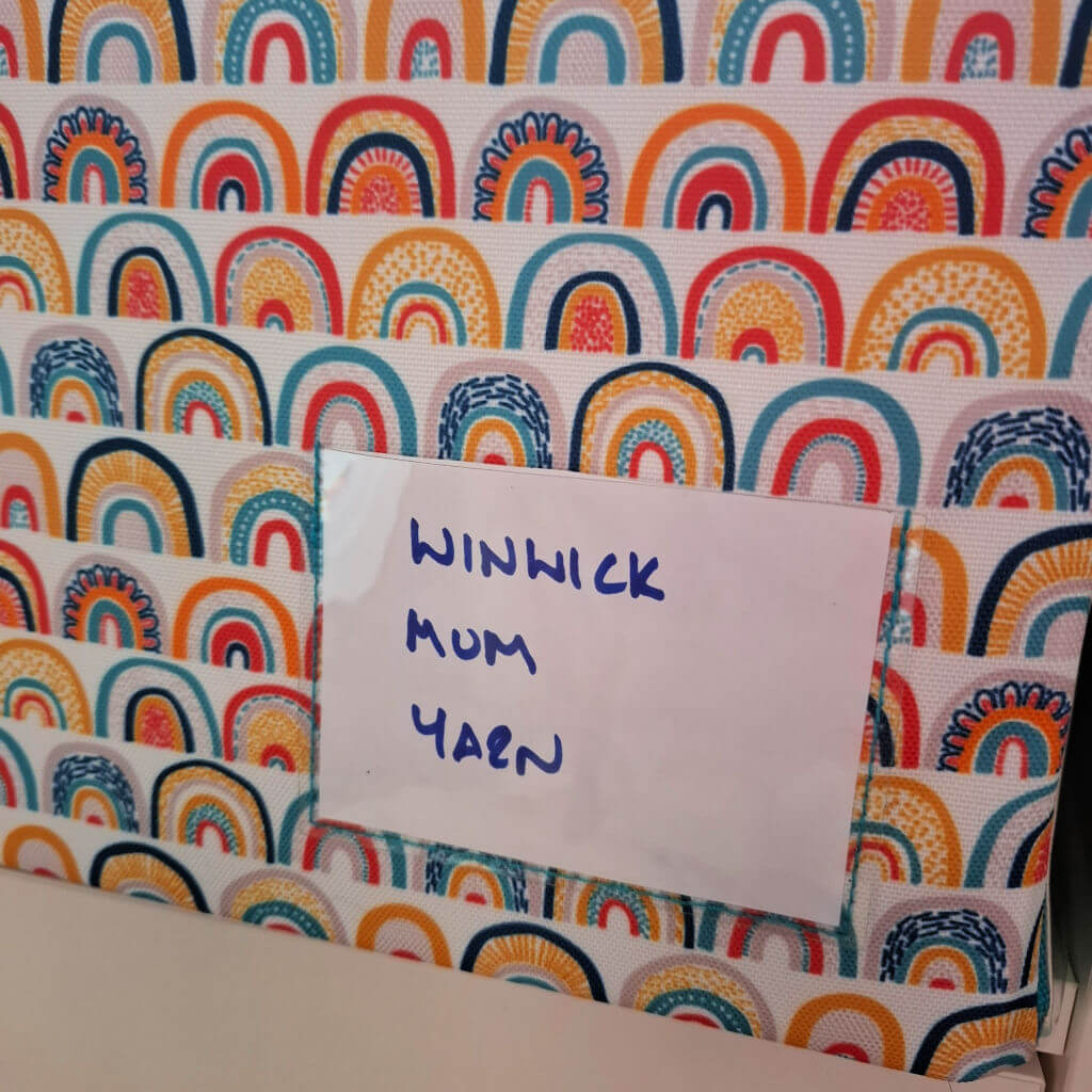 Colourful fabric featuring rainbow patterns with a white label that reads "WINWICK MUM YARN" in blue handwriting. The tone is cheerful and vibrant.