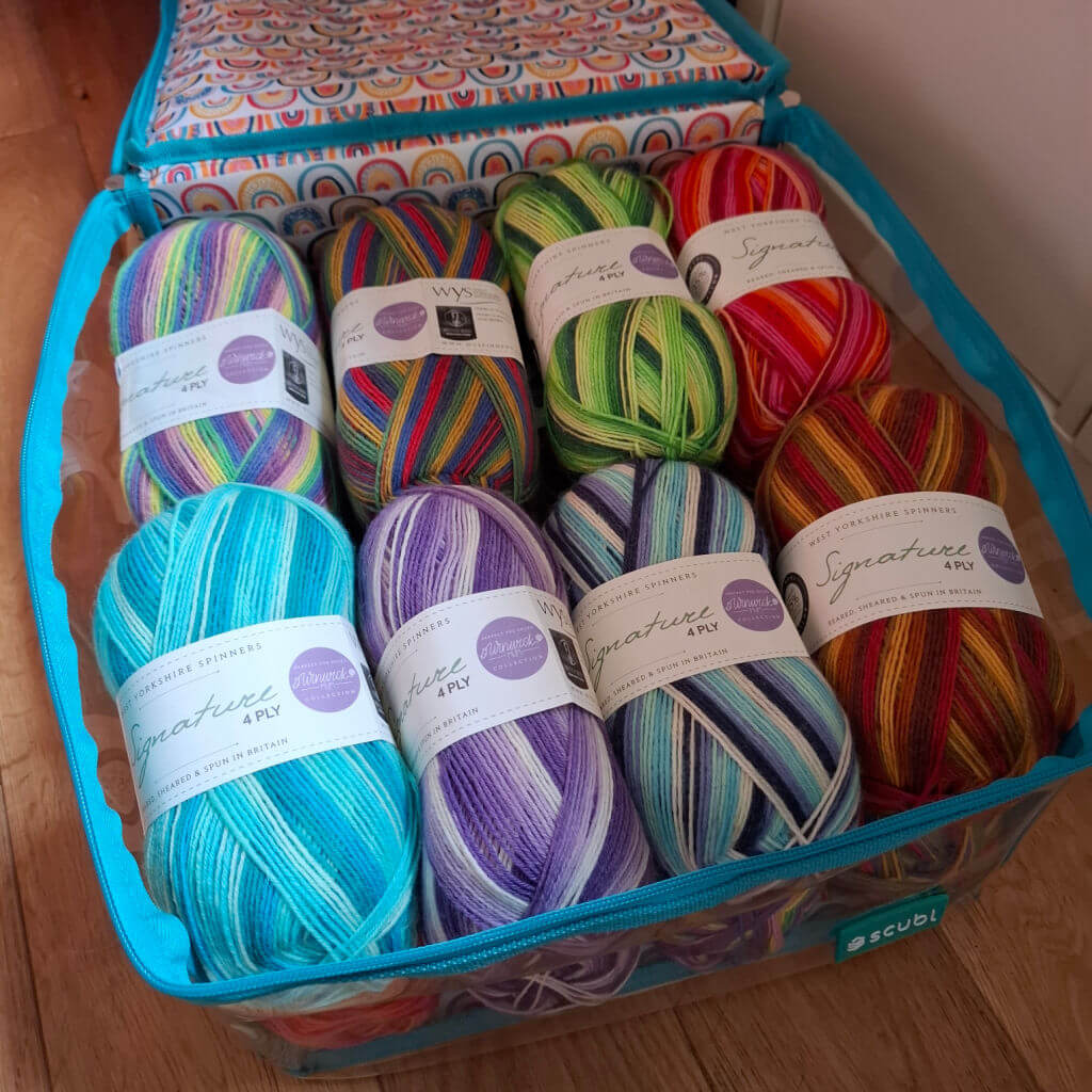 A colourful assortment of Winwick Mum yarn balls arranged neatly in a blue storage bag. The yarn features vibrant stripes in blues, greens, purples, and reds, exuding a creative and lively tone.