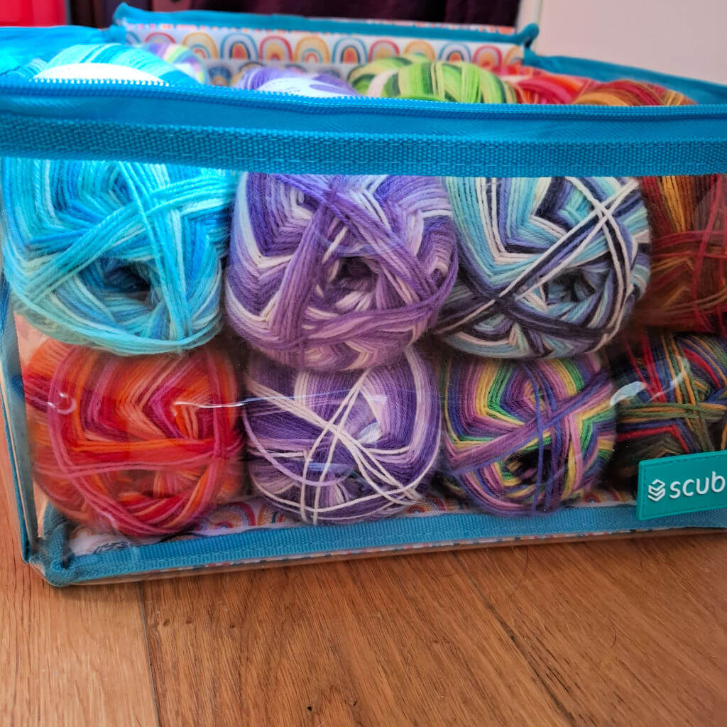 Transparent Scubl box filled with colourful yarn balls in blues, purples, and reds, on a wooden floor. The setup conveys a cheerful, crafty tone.