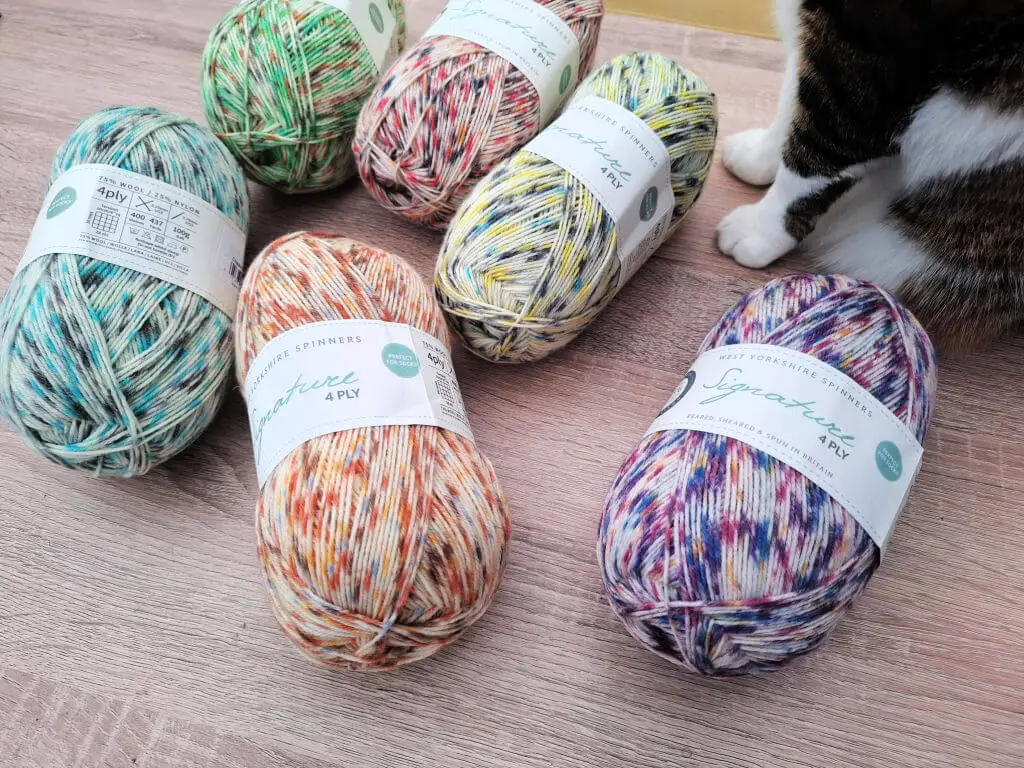 Six colourful skeins of Close-up of West Yorkshire Spinners Signature 4ply Butterflies collection yarn are arranged on a wooden surface, featuring vibrant hues of blue, green, orange, yellow, and purple. A cat's paws are visible nearby, adding a cozy touch.