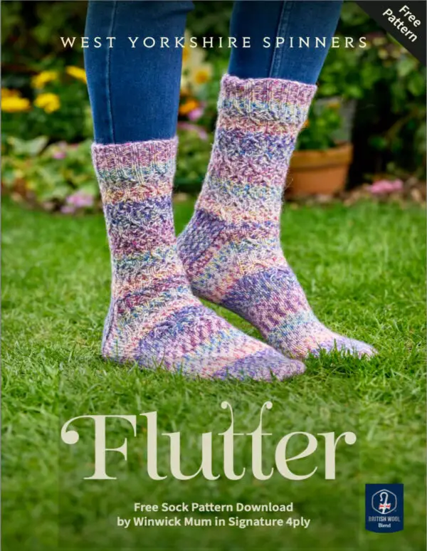 Colourful knitted socks with a textured pattern, worn by someone in jeans, standing on lush green grass in a garden with flowers in the background. Winwick Mum pattern, West Yorkshire Spinners Flutter