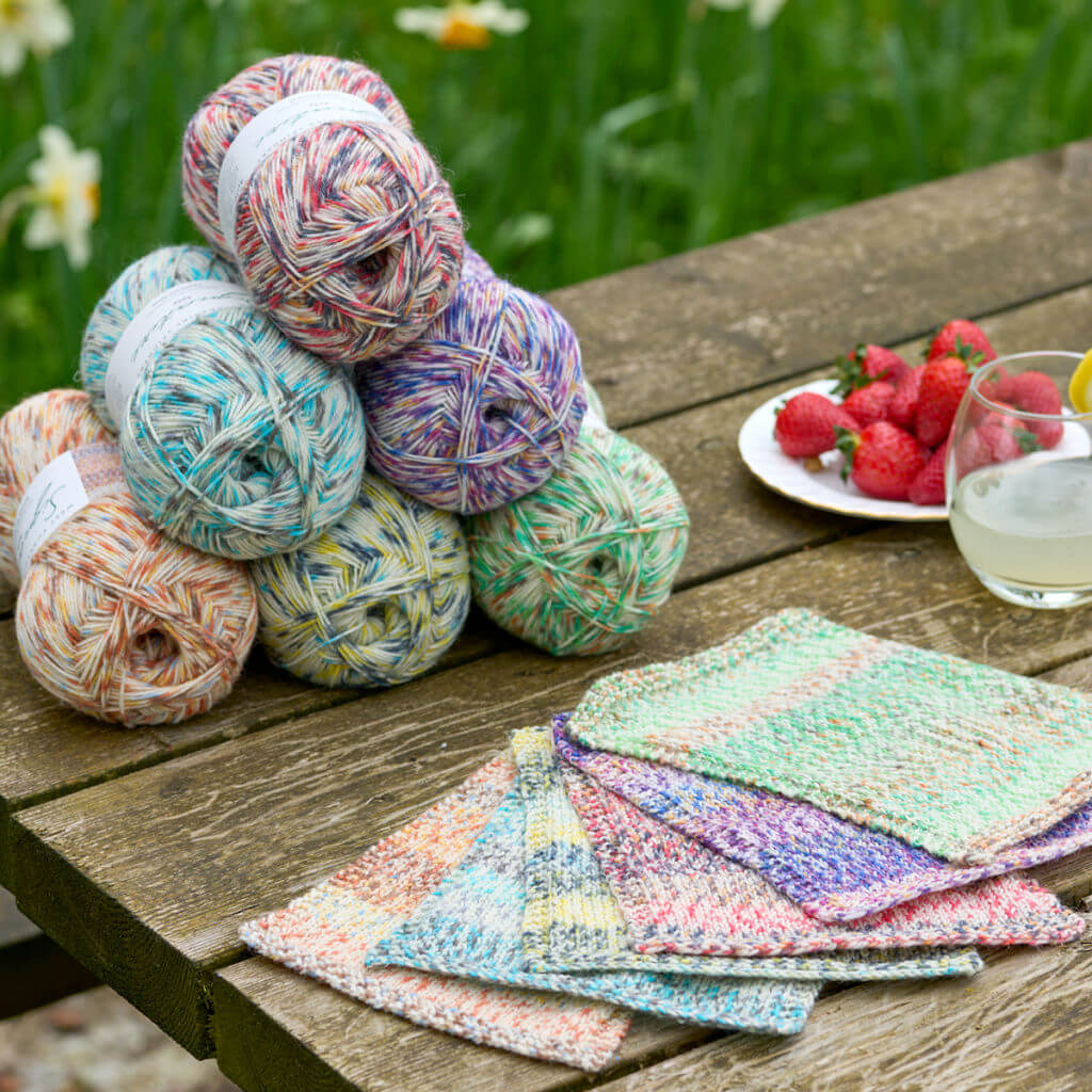 Stacked yarn balls in various pastel shades sit beside colourful knitted squares on a wooden table. A plate of strawberries and a glass of lemonade add a fresh, summery vibe. West Yorkshire Spinners Butterflies yarn