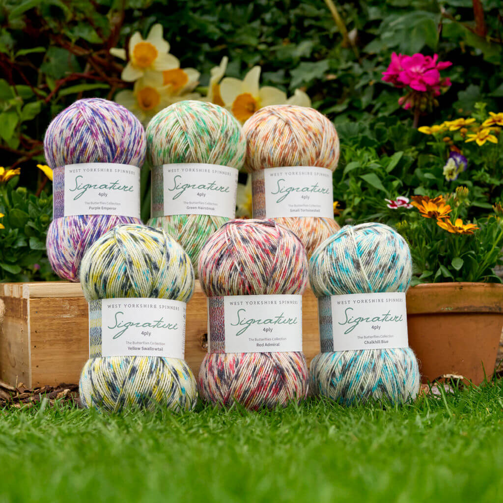 Balls of colourful West Yorkshire Spinners Butterflies yarn in purple, green, orange, yellow, red, and blue are arranged on a wooden crate. A vibrant garden with flowers forms the backdrop.