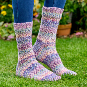 Colourful knitted socks with a textured pattern, worn by someone in jeans, standing on lush green grass in a garden with flowers in the background. Winwick Mum pattern, West Yorkshire Spinners Flutter