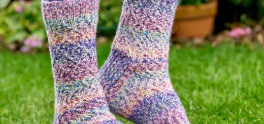 Colourful knitted socks with a textured pattern, worn by someone in jeans, standing on lush green grass in a garden with flowers in the background. Winwick Mum pattern, West Yorkshire Spinners Flutter