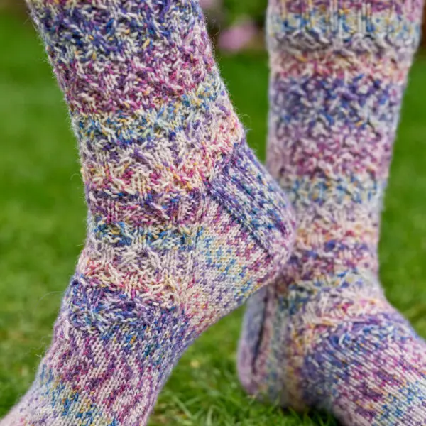 Close-up of colourful, textured knit socks in pastel shades of purple, blue, and yellow. They rest on a green grassy background, conveying warmth and coziness.