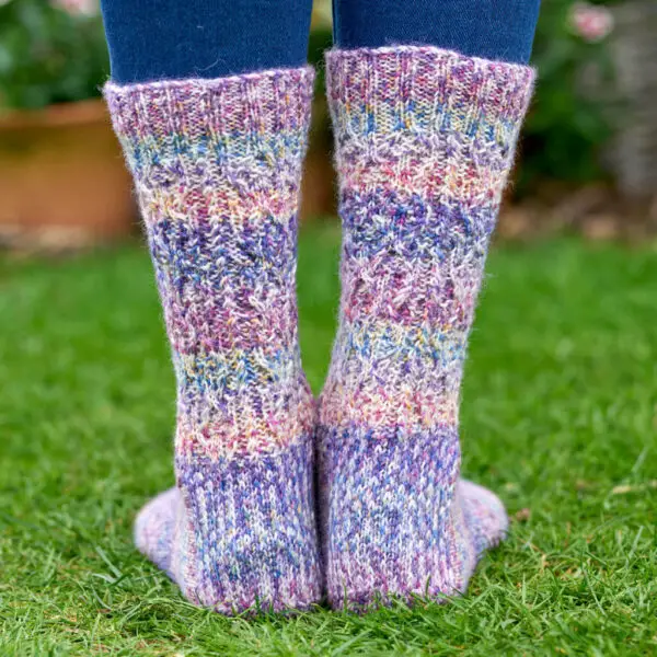 Colourful knitted socks with a purple, pink, and blue pattern, worn over jeans, standing on green grass. The tone is warm and cozy.
