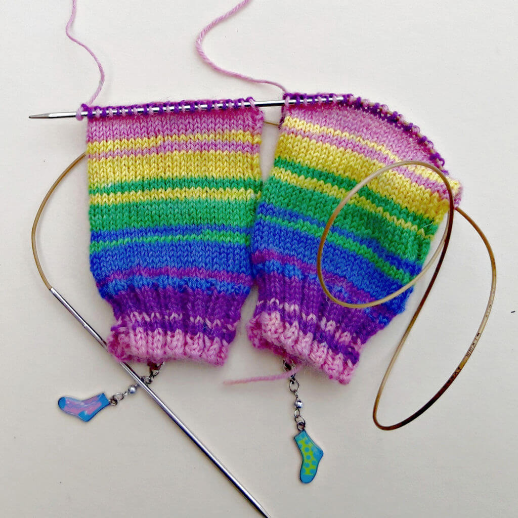 Two colourful knitted pieces on one circular knitting needle featuring vibrant stripes of purple, pink, yellow, green, and blue, with sock-shaped stitch markers. Winwick Mum | Basic 4ply Socks | Beginner sock knitting | Free sock knitting patterns | TAAT two at a time socks