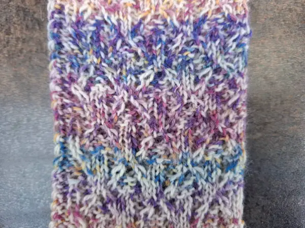 Close-up of a knitted fabric with a textured zig-zag pattern. The yarn is multicolored, featuring shades of purple, blue, and white, creating a cozy, intricate design.