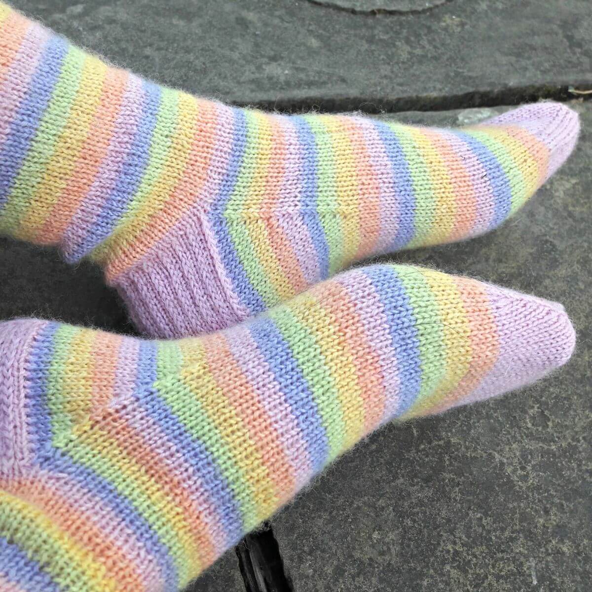 Close-up of two feet wearing pastel-striped socks in shades of purple, yellow, blue, green, and pink. The socks rest on grey stone paving flags. Winwick Mum | Basic 4ply Socks | Beginner sock knitting | Free sock knitting patterns | Contrast cuffs, heels and toes