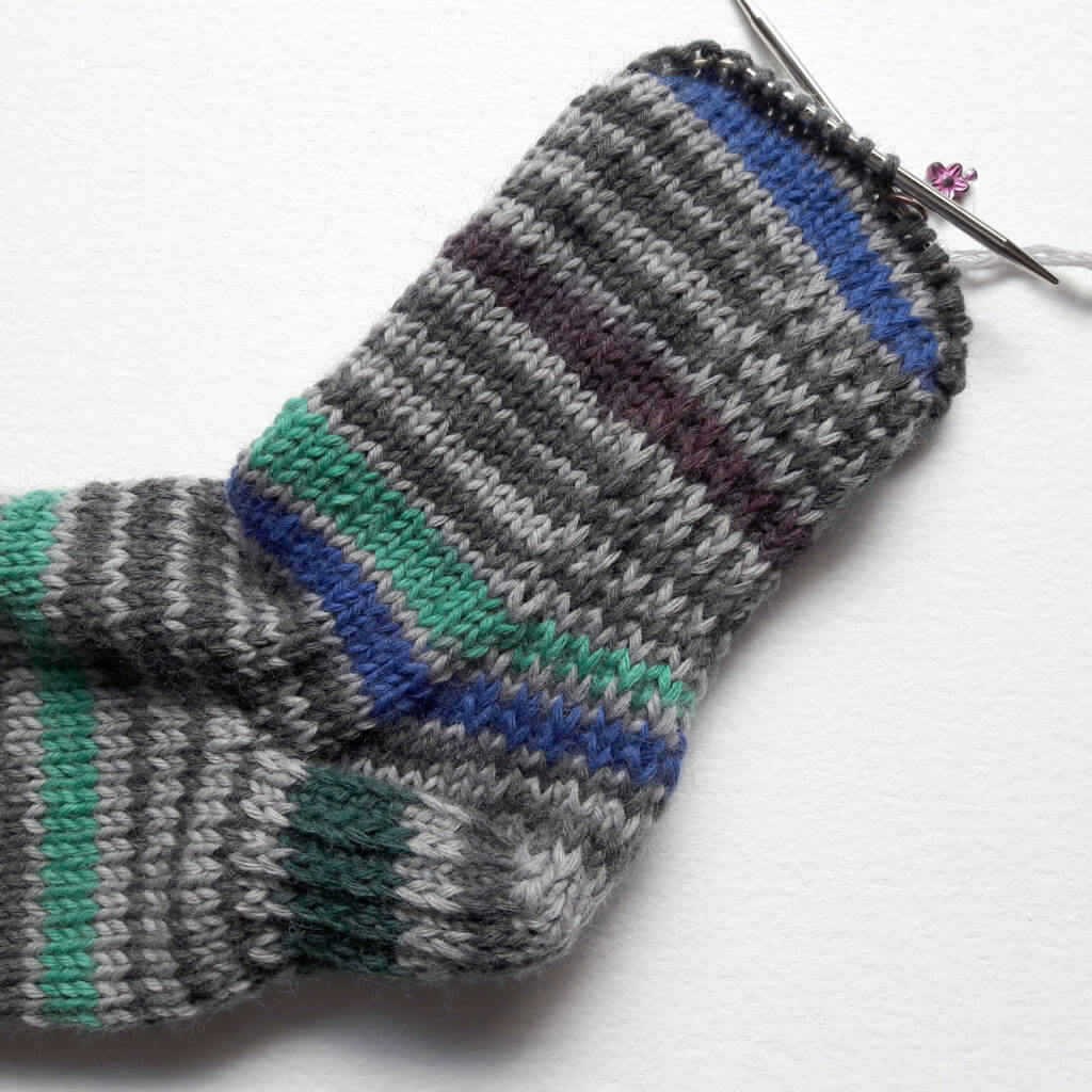 Knitted sock in progress on knitting needles, featuring horizontal stripes in gray, black, blue, and green. Background is a soft white surface. Winwick Mum | Beginner sock knitting | Free sock knitting patterns | Reinforcing sock heels, soles and toes
