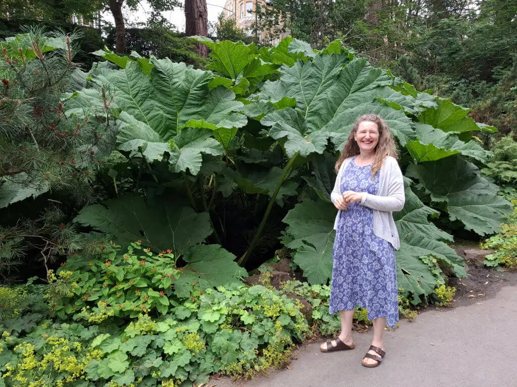 A woman in a purple patterned dress and grey cardigan stands smiling in front of large green leaves in a lush garden, conveying a sense of joy.