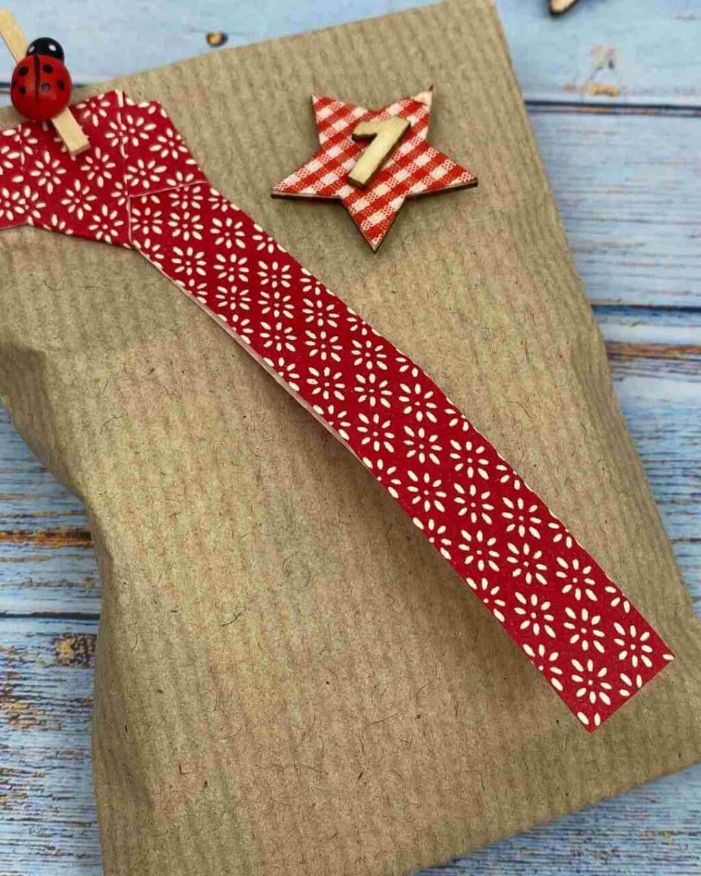 A brown paper package is adorned with red patterned tape, a checkered star marked "1," and a small ladybird clip, resting on a textured blue wooden surface.