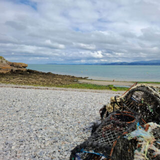 Pebble beach with a pile of fishing nets in the foreground. The calm sea stretches to the horizon under a cloudy sky, creating a serene atmosphere.