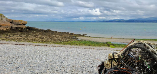Pebble beach with a pile of fishing nets in the foreground. The calm sea stretches to the horizon under a cloudy sky, creating a serene atmosphere.