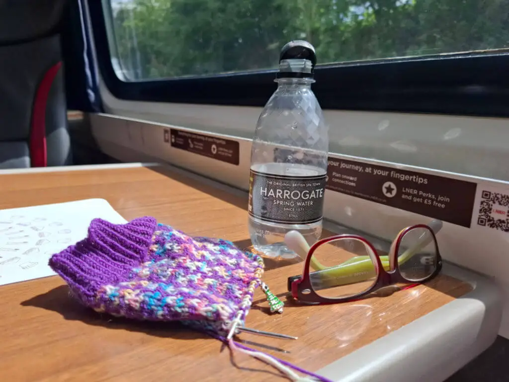 Knitting project with purple and multicoloured yarn, glasses, and a Harrogate water bottle on a train table by a window overlooking greenery. Cosy and creative.