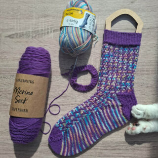 A colourful knitted sock is displayed on a light wooden surface. Nearby are a purple yarn ball and multicoloured yarn. A cat's paw touches the sock.