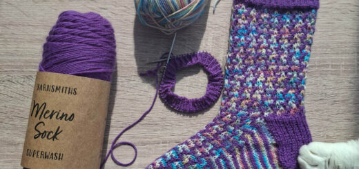 A colourful knitted sock is displayed on a light wooden surface. Nearby are a purple yarn ball and multicoloured yarn. A cat's paw touches the sock.
