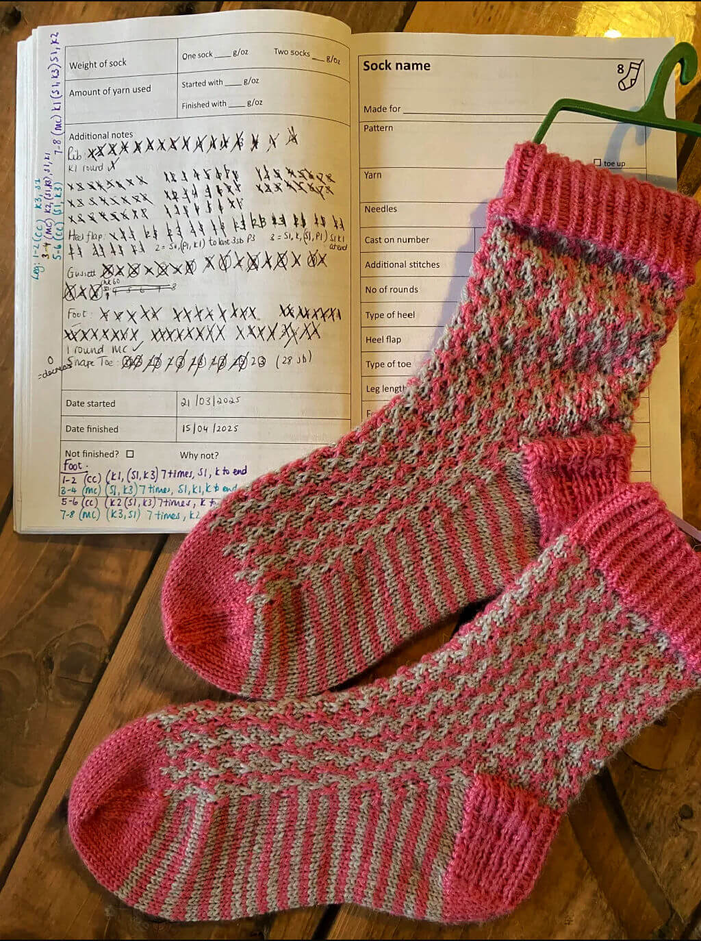 Knitted pink and gray socks with a textured pattern lay on an open notebook with handwritten knitting notes. The wooden background adds warmth.