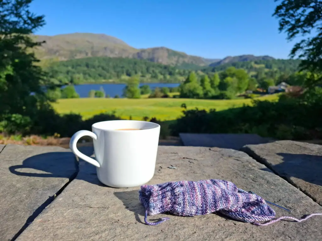 A white mug of tea and a small knitting project rest on a stone table, set against a lush green landscape with a lake and mountains under a clear blue sky.