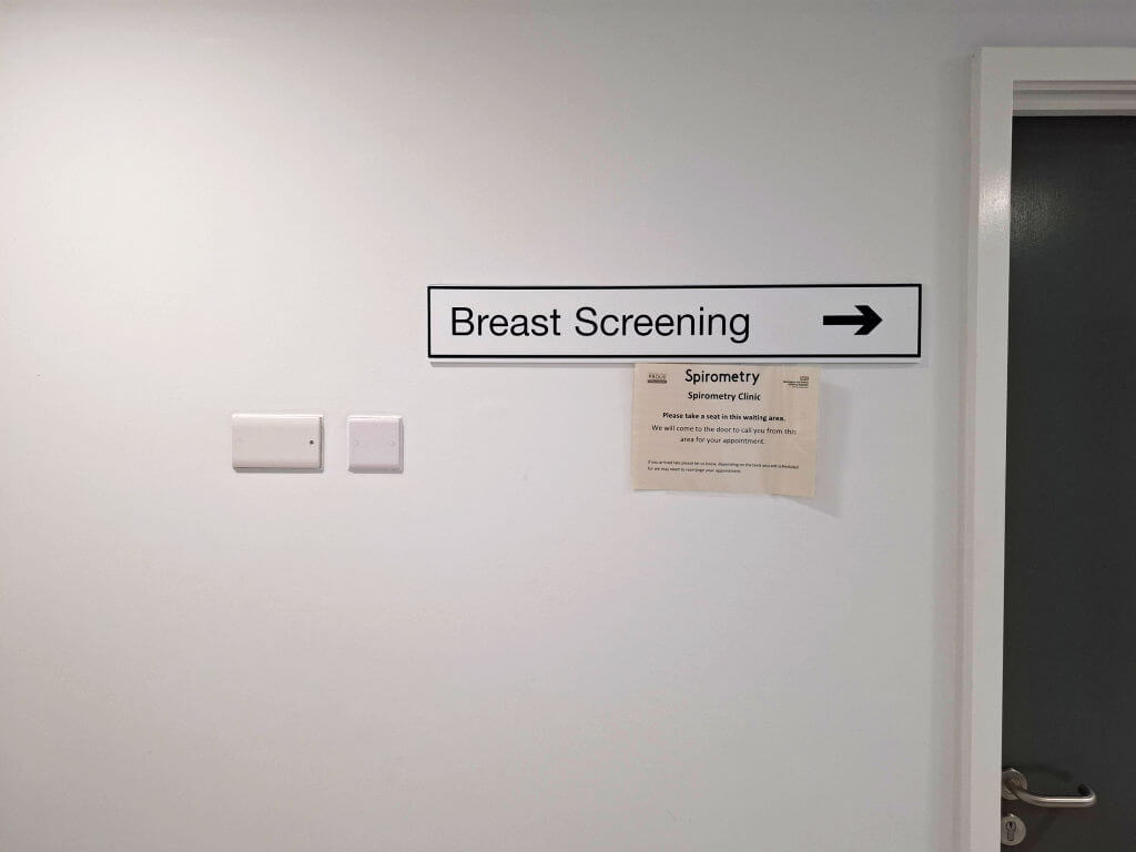 A sign on a white wall points right to "Breast Screening." Below, a smaller notice about the Spirometry Clinic is posted. Nearby, a closed brown door.