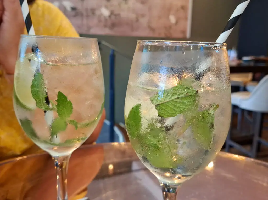 Two glasses filled with a clear, fizzy drink, garnished with fresh mint leaves and lime slices, have striped straws. A relaxed, cosy restaurant setting.