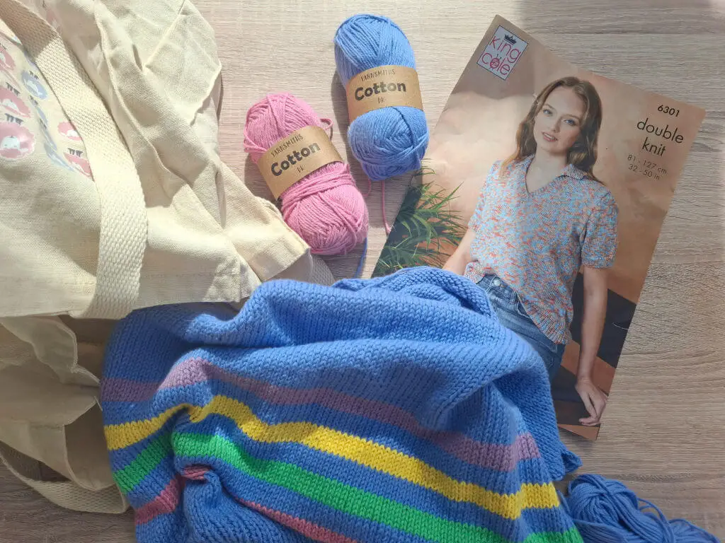 Craft supplies on a wooden table, featuring pastel cotton yarn, a knitting pattern booklet, a colourful striped knit piece, and a tote bag.