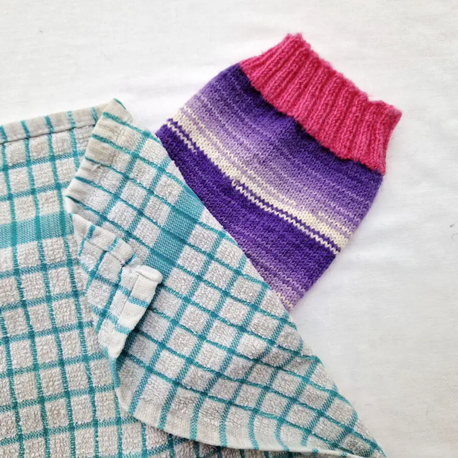 A single purple and white sock with pink cuffs, heels and toes lie on a white background. The sock is party covered by a blue and white checked tea towel. Winwick Mum | Blocking socks with sock blockers