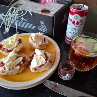 Four half scones with cream and jam on a yellow plate next to a cut glass containing Pimm's and fruit. The empty Pimm's can is next to the glass.