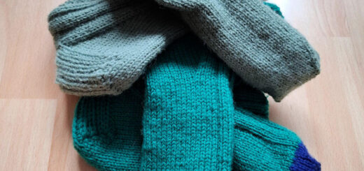 A pile of four knitted socks on a wooden floor. Two are dark green with blue toes, and two are sage green. The socks appear cosy and handmade.
