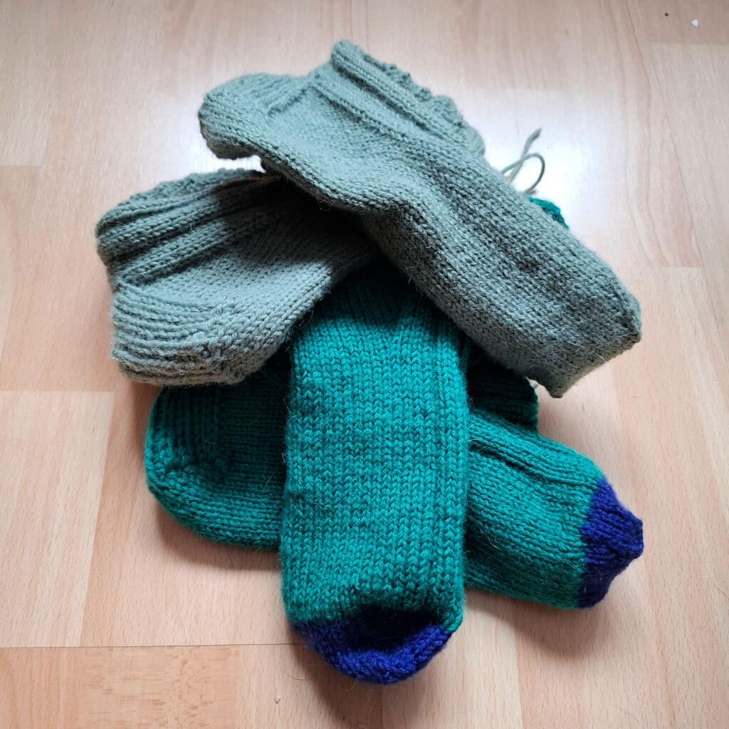 A pile of four knitted socks on a wooden floor. Two are dark green with blue toes, and two are sage green. The socks appear cosy and handmade.