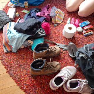 Clothes, hiking boots, trainers, backpack, toiletries, snacks, and a roll of toilet paper are spread on a red rug, suggesting a packing scene.