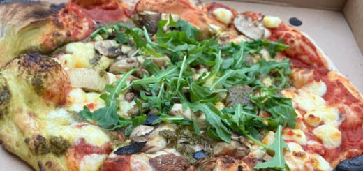 Wood-fired pizza topped with rocket, mushrooms, and cheese, showcasing charred crust in an open box. Background features colourful beer cans.