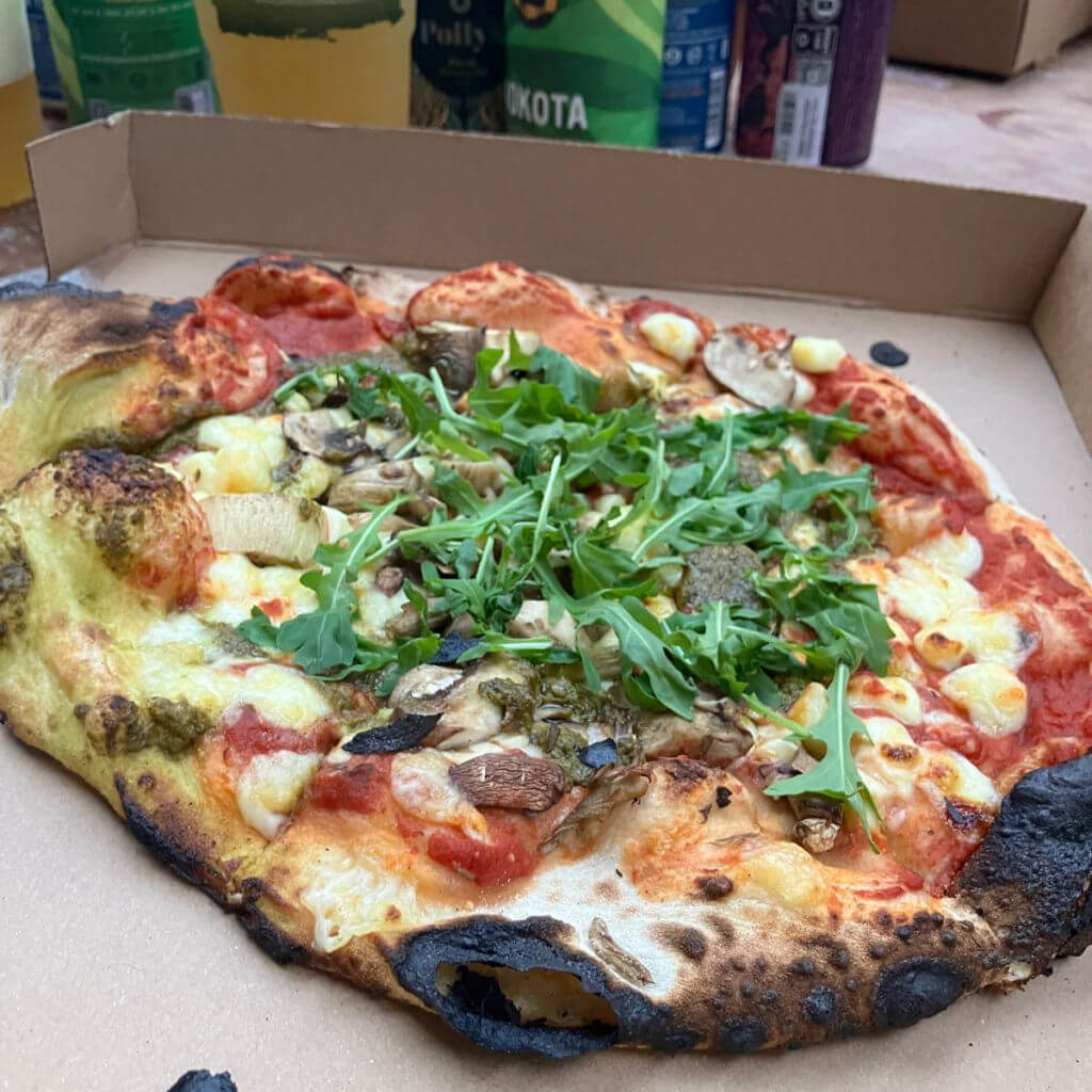 Wood-fired pizza topped with rocket, mushrooms, and cheese, showcasing charred crust in an open box. Background features colourful beer cans.
